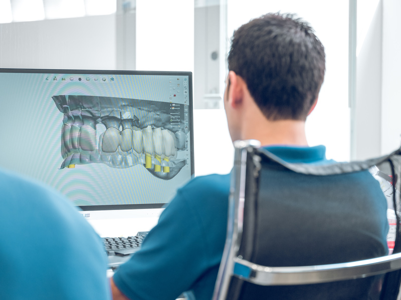 Back view of unrecognizable dental technician sitting on chair in modern clinic and looking at digital 3d model of teeth on display